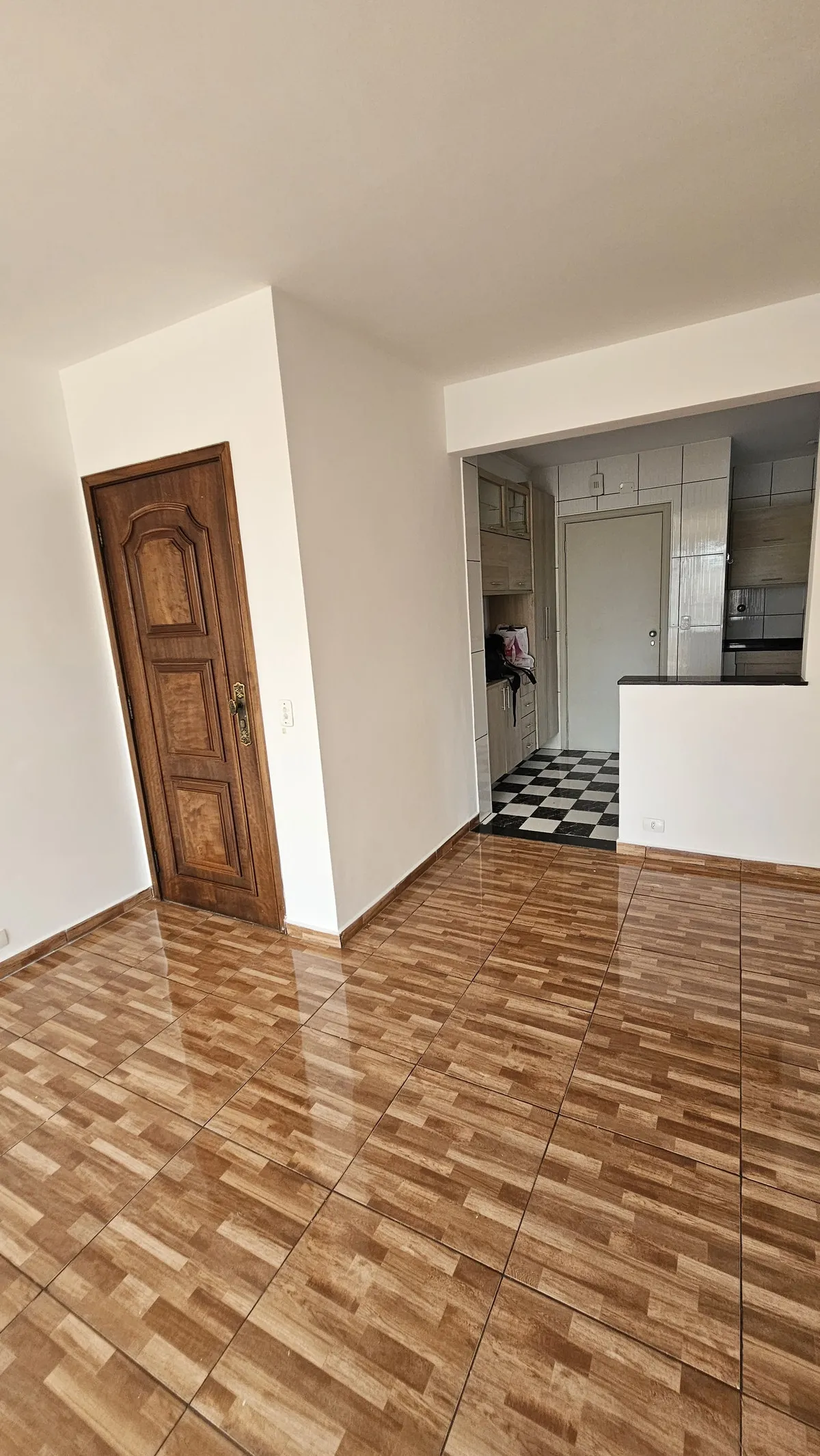 Sala com porta de madeira antiga entalhada, piso cerâmico amadeirado e cozinha de piso xadrez ao fundo, antes da reforma