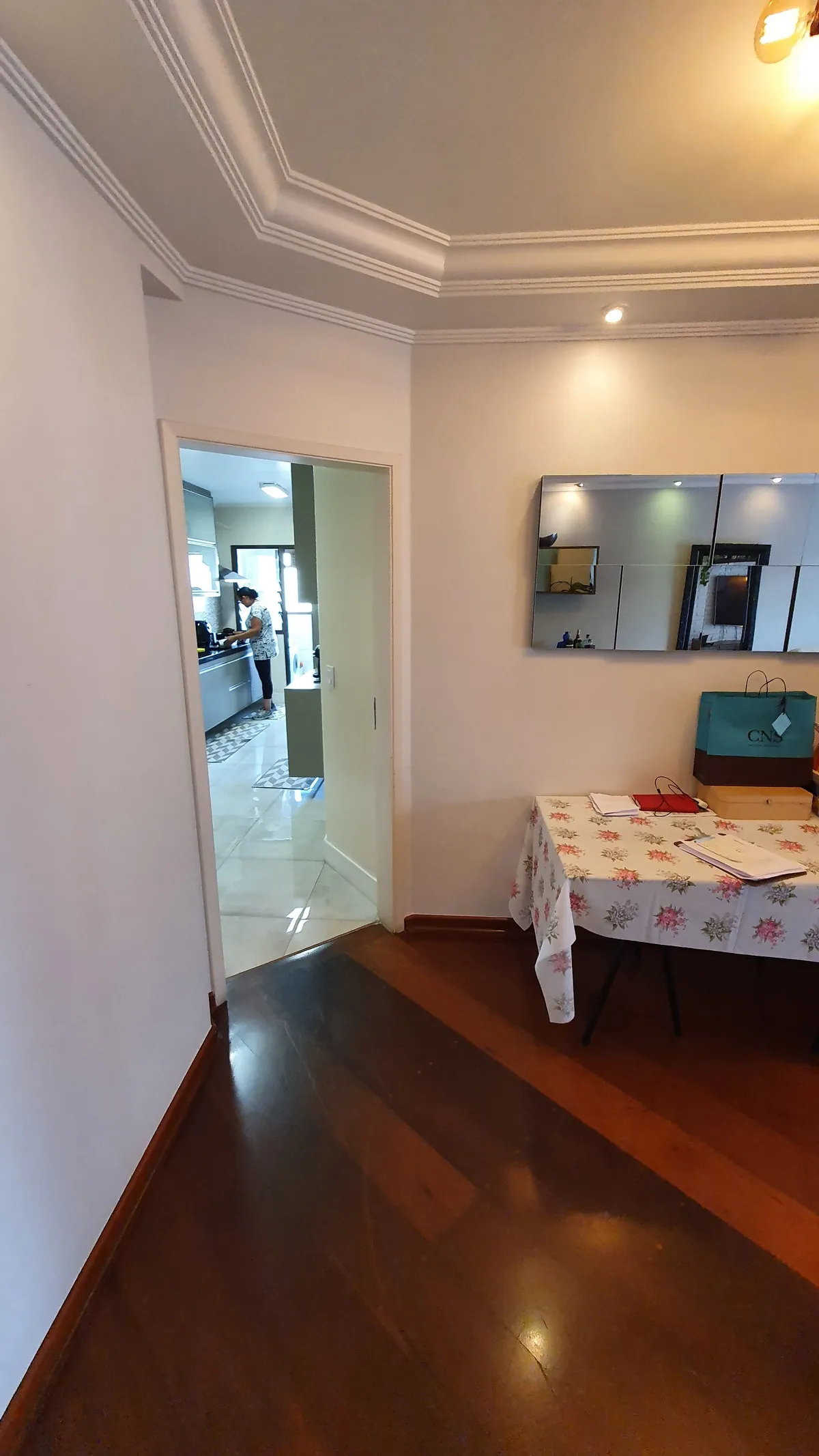 Corredor com piso de madeira escura, mesa de toalha floral e painel de espelhos na parede, antes da reforma