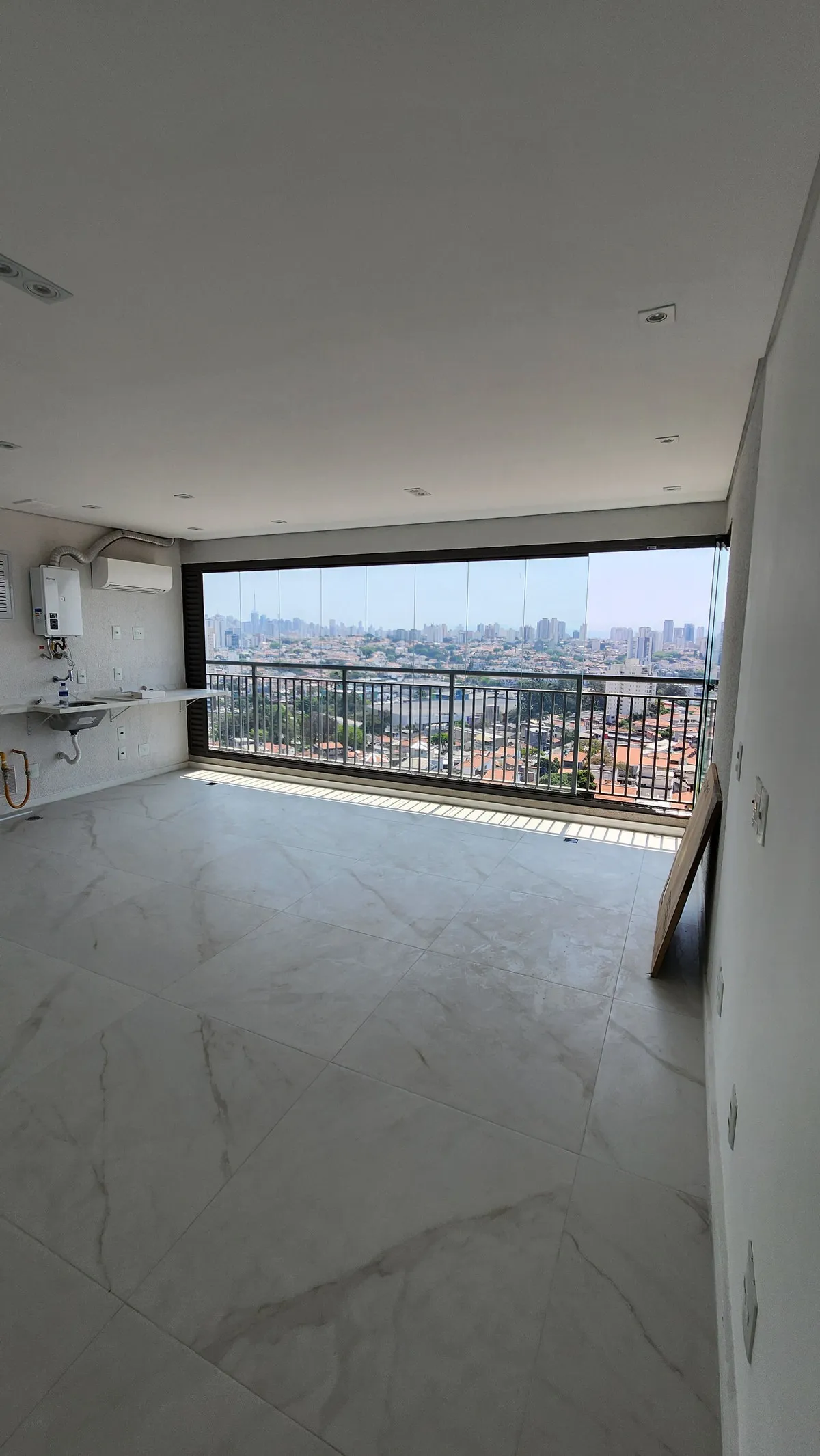 Living vazio com varanda envidraçada e vista da cidade, aquecedor a gás e pia na parede, antes da reforma