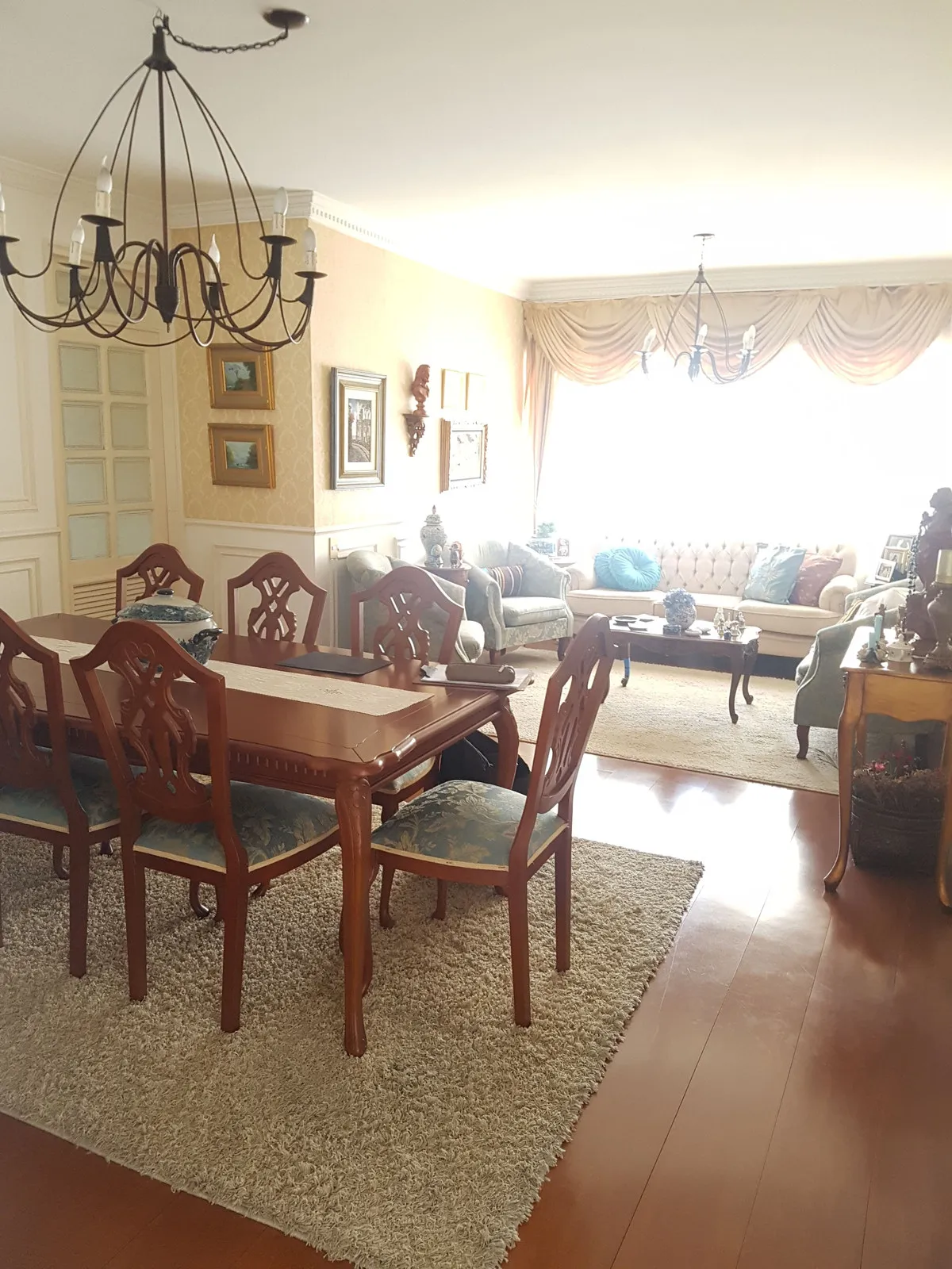 Sala de jantar e estar antigas, com mesa e cadeiras de madeira escura entalhada, tapete bege e lustre de ferro, antes da reforma