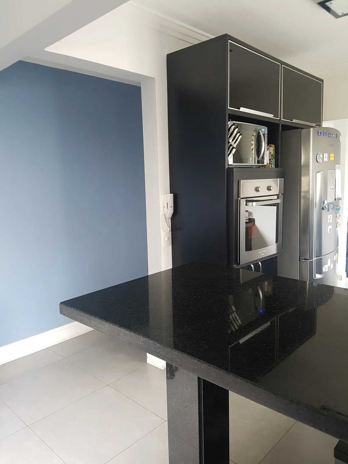 Cozinha antiga com parede azul, bancada de granito preto, torre de armários pretos com forno e geladeira inox, antes da reforma