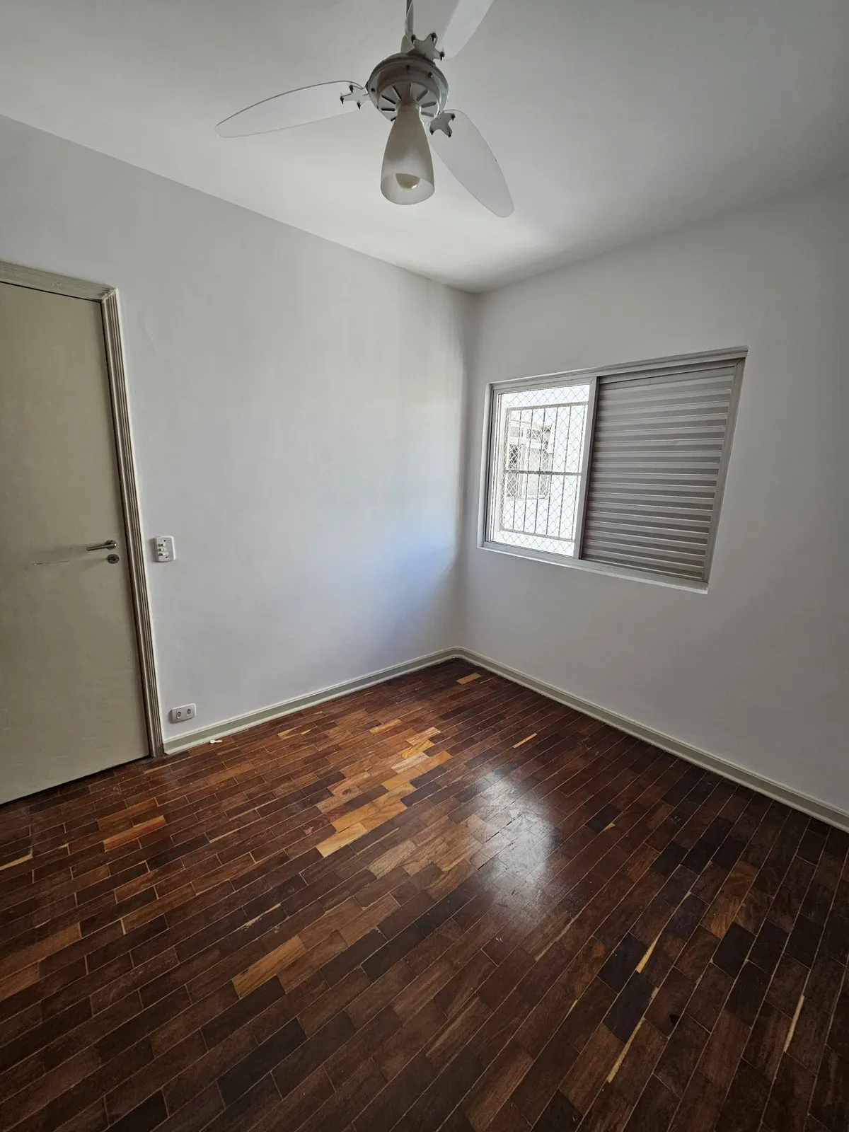 Quarto vazio com paredes claras, piso de taco escuro, janela com persiana e ventilador, antes da reforma
