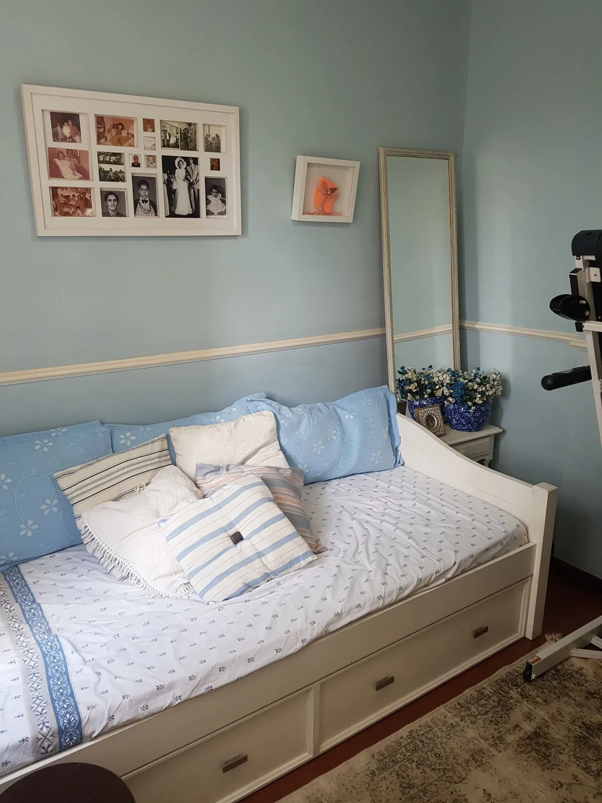 Quarto azul-claro com cama branca, roupa de cama azul, espelho de chão e quadros de família, antes da reforma
