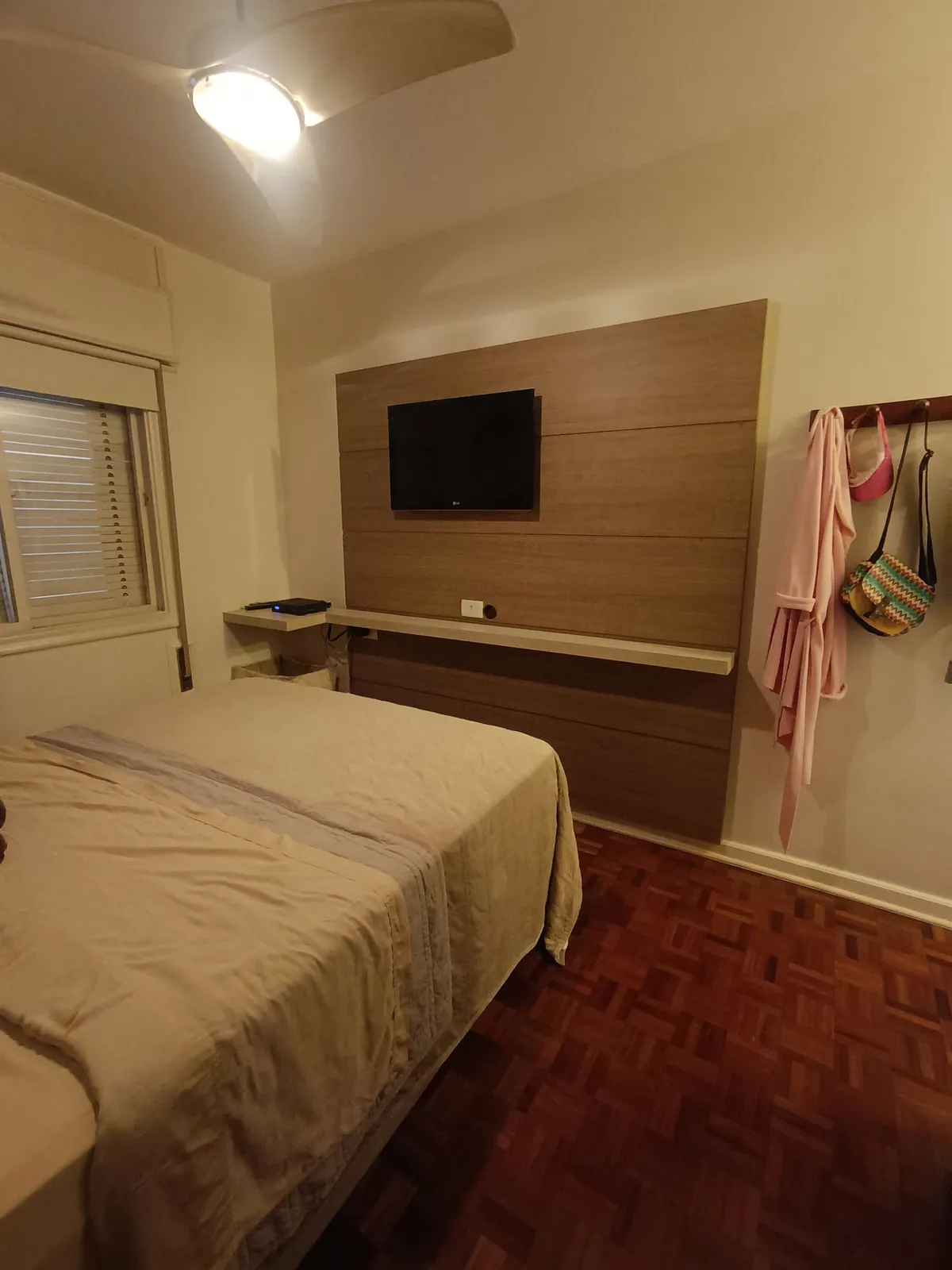 Quarto de casal com painel de madeira, TV, cabideiro na parede e piso de taco, antes da reforma