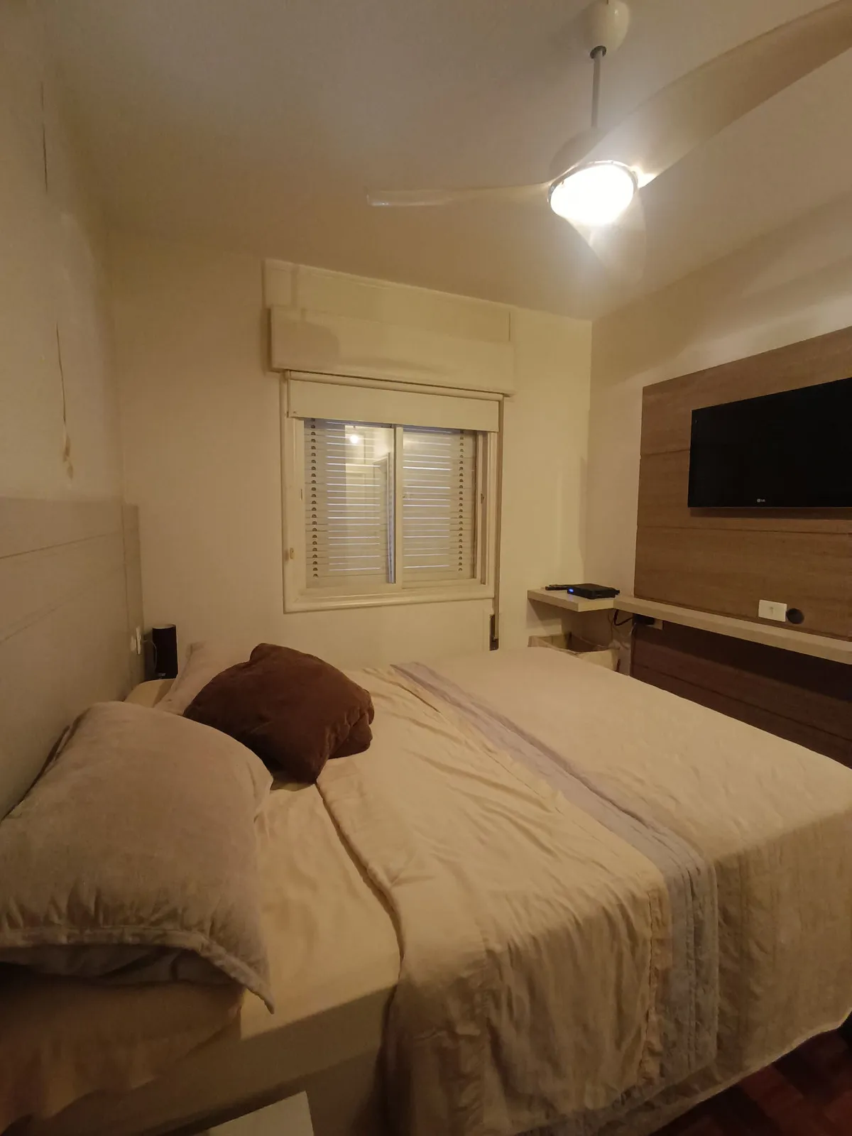 Quarto de casal com painel de madeira, TV, janela com persiana e piso de taco, antes da reforma