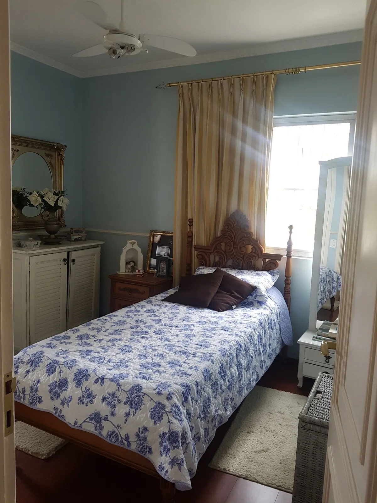 Quarto azul com cabeceira de madeira entalhada, colcha floral azul e cortina dourada, antes da reforma