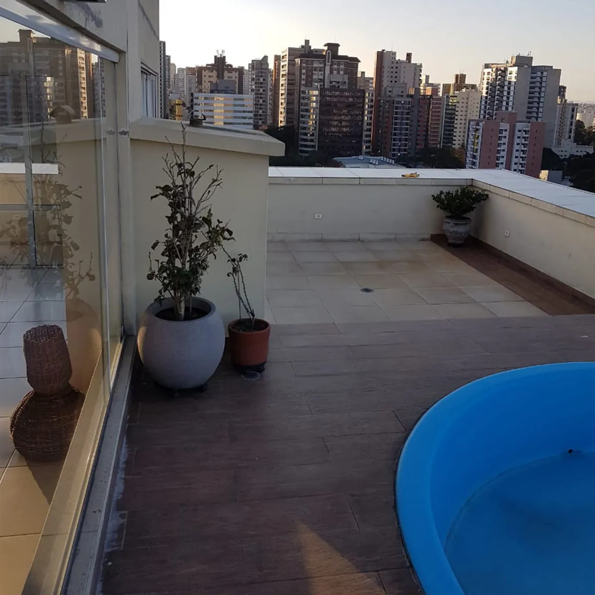 Terraço da cobertura com deck de madeira, vasos de plantas e piscina azul ao entardecer, antes da reforma