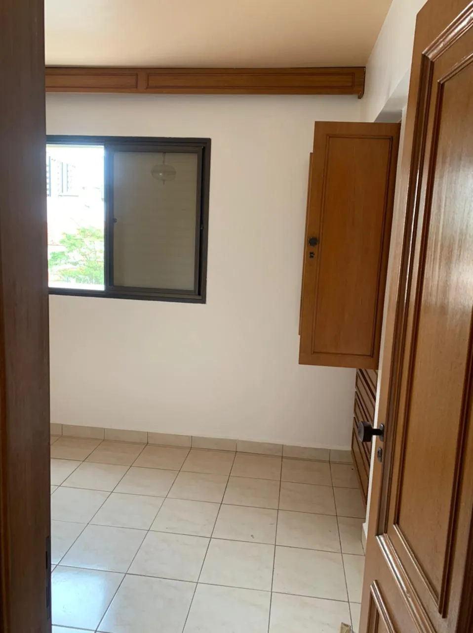 Quarto vazio com bandô de madeira, janela, armário embutido de madeira na parede e piso de cerâmica, antes da reforma