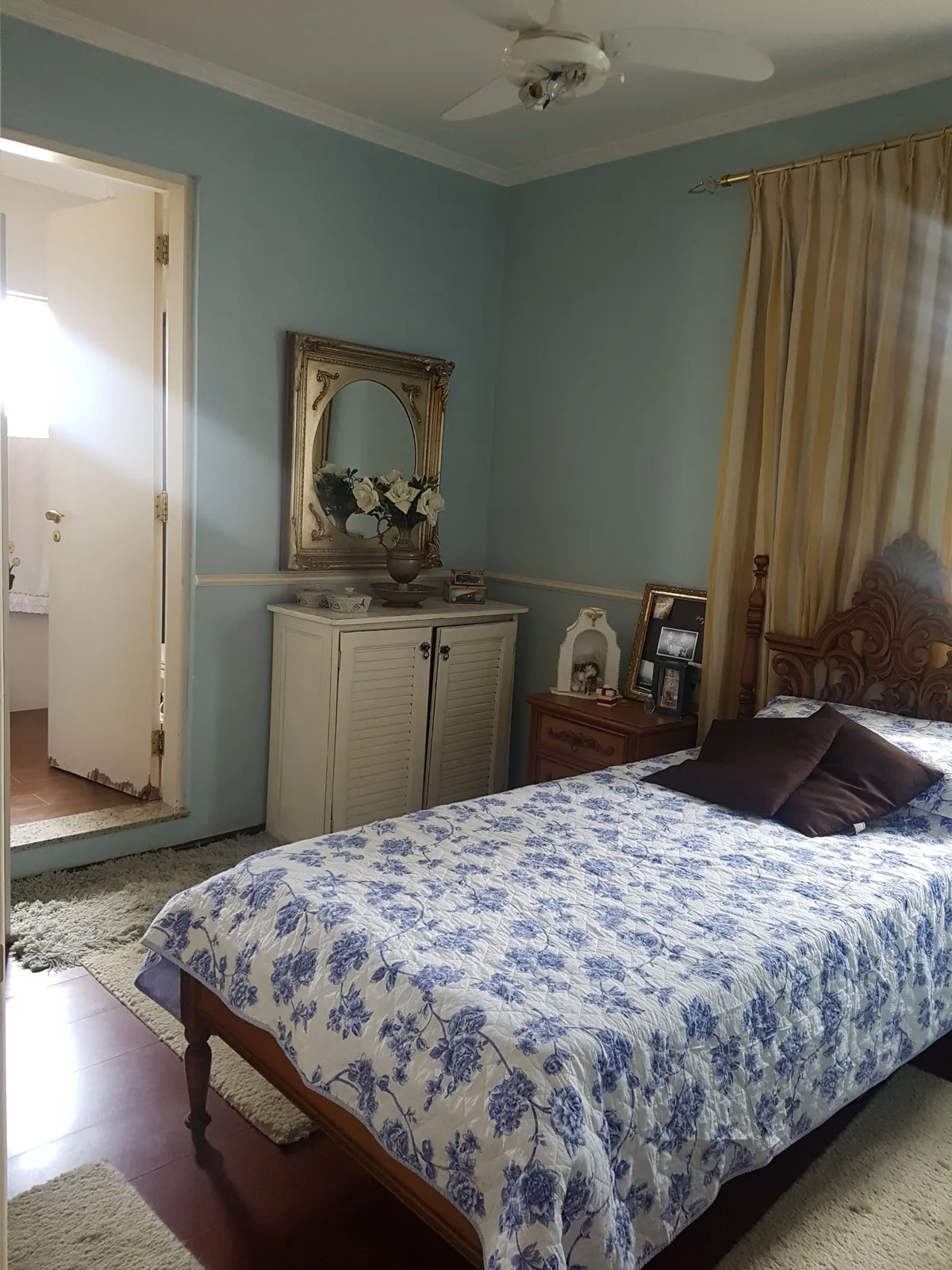 Quarto azul com cômoda branca, espelho dourado ornamentado e cama de colcha floral azul, antes da reforma