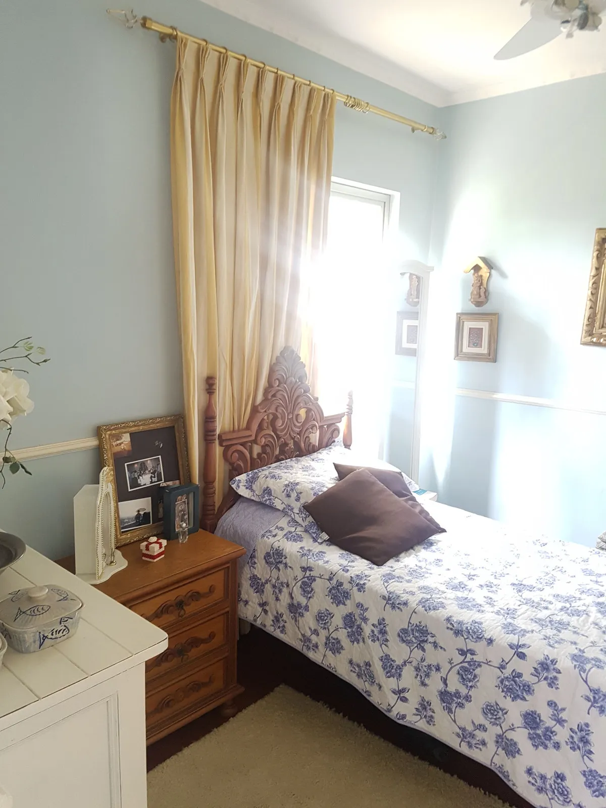 Quarto azul com criado-mudo de madeira, colcha floral azul e cabeceira entalhada, antes da reforma