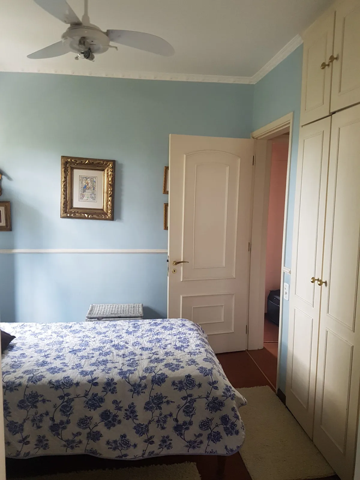 Quarto azul com cama de colcha floral, porta branca e armário off-white embutido, antes da reforma