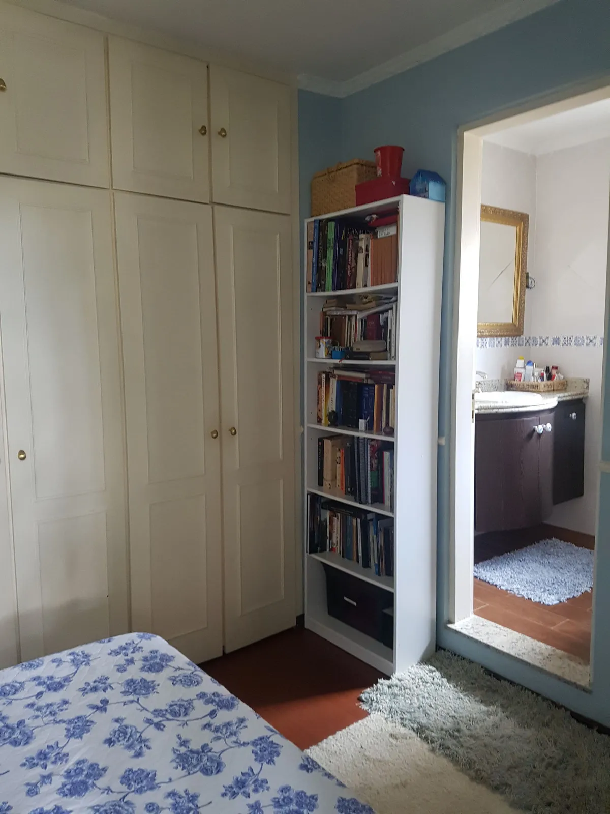 Quarto azul com armário off-white, estante de livros e banheiro de gabinete escuro ao fundo, antes da reforma