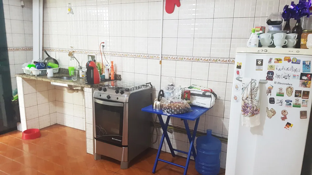 Cozinha antiga com bancada de granito, fogão de inox, mesa dobrável azul e geladeira cheia de adesivos, antes da reforma
