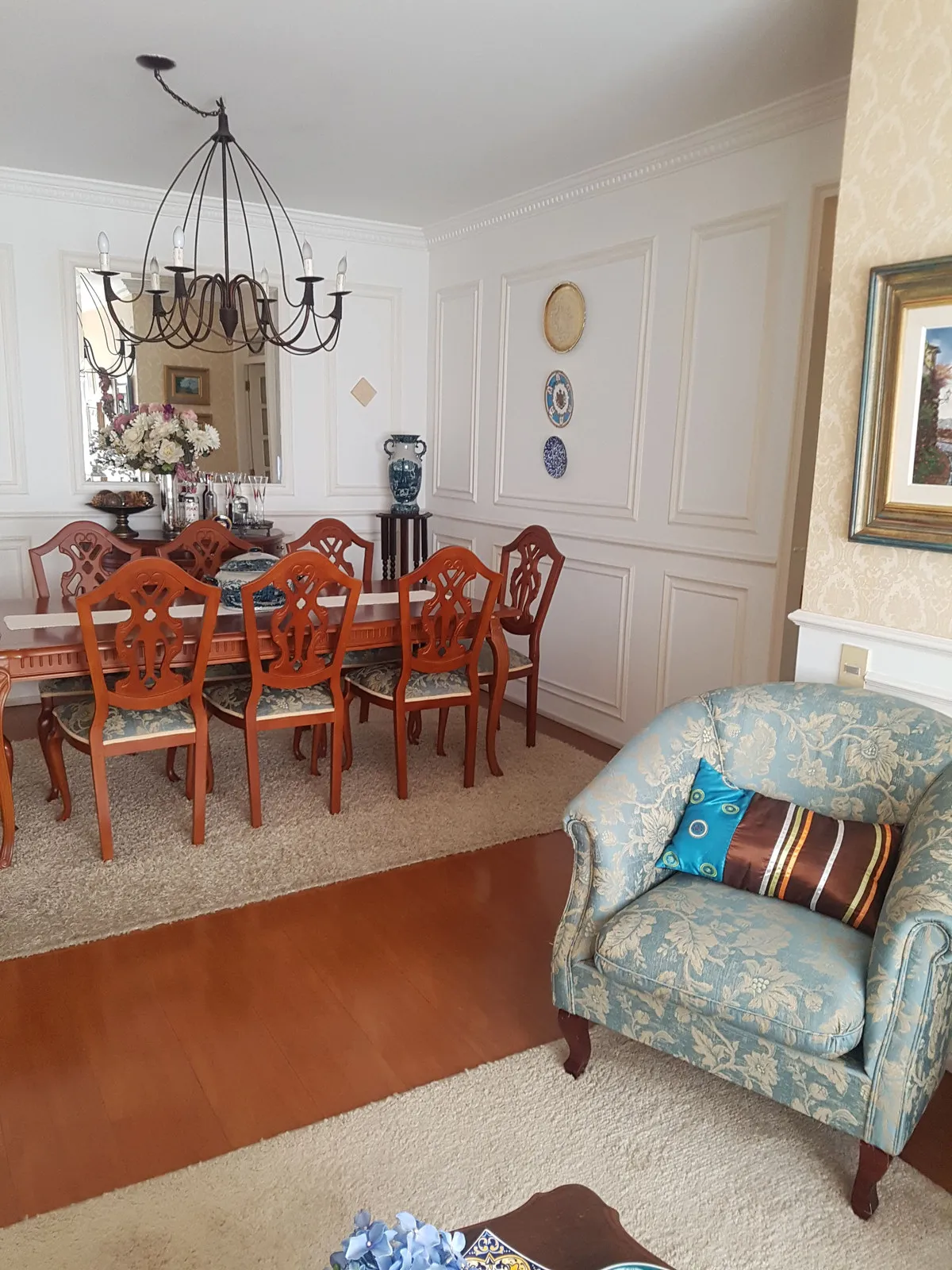 Sala de jantar com cadeiras de madeira escura, boiserie branca, poltrona floral azul e piso de madeira alaranjado, antes da reforma
