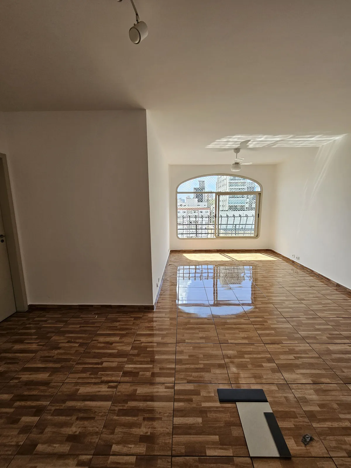 Sala ampla vazia com piso cerâmico marrom brilhante, janela em arco e trilho de spots no teto, antes da reforma