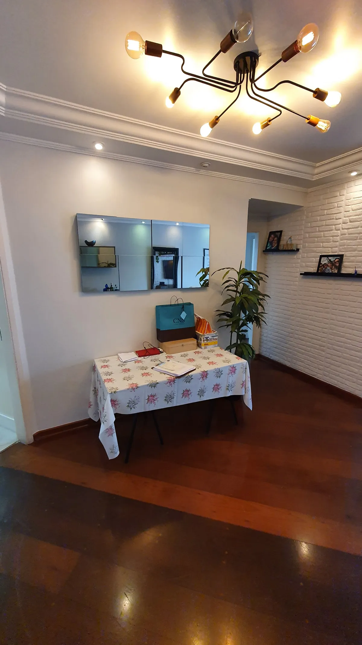 Sala de jantar com mesa de toalha floral, painel de espelhos e parede de tijolo branco, antes da reforma