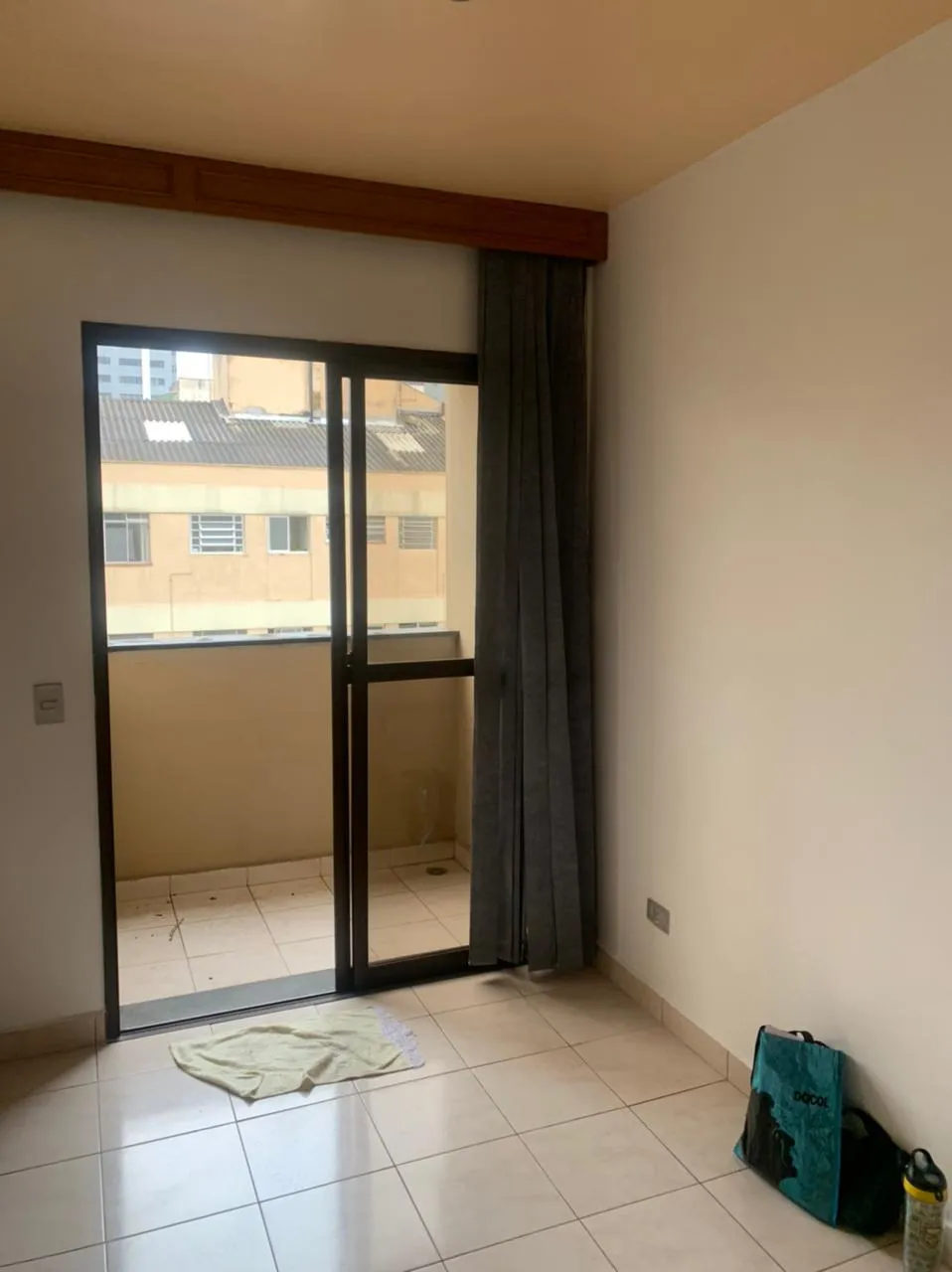 Sala vazia com porta de correr para a varanda, cortina cinza e piso de cerâmica, antes da reforma