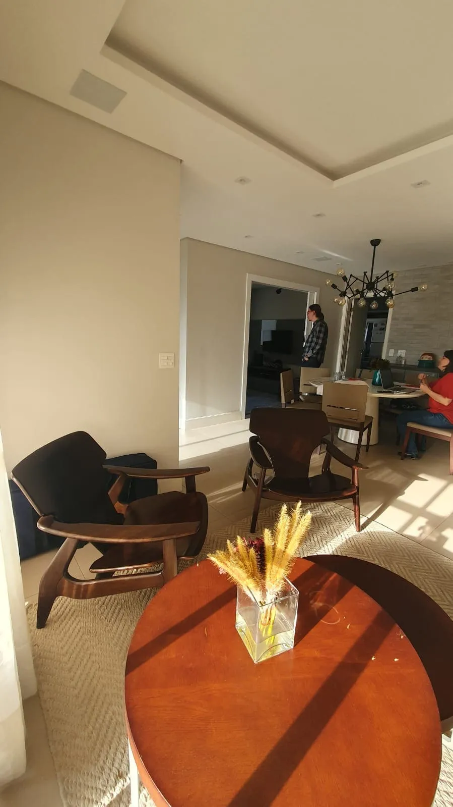 Estar antigo com poltronas de madeira escura, mesa de centro redonda e sala de jantar ao fundo, antes da reforma