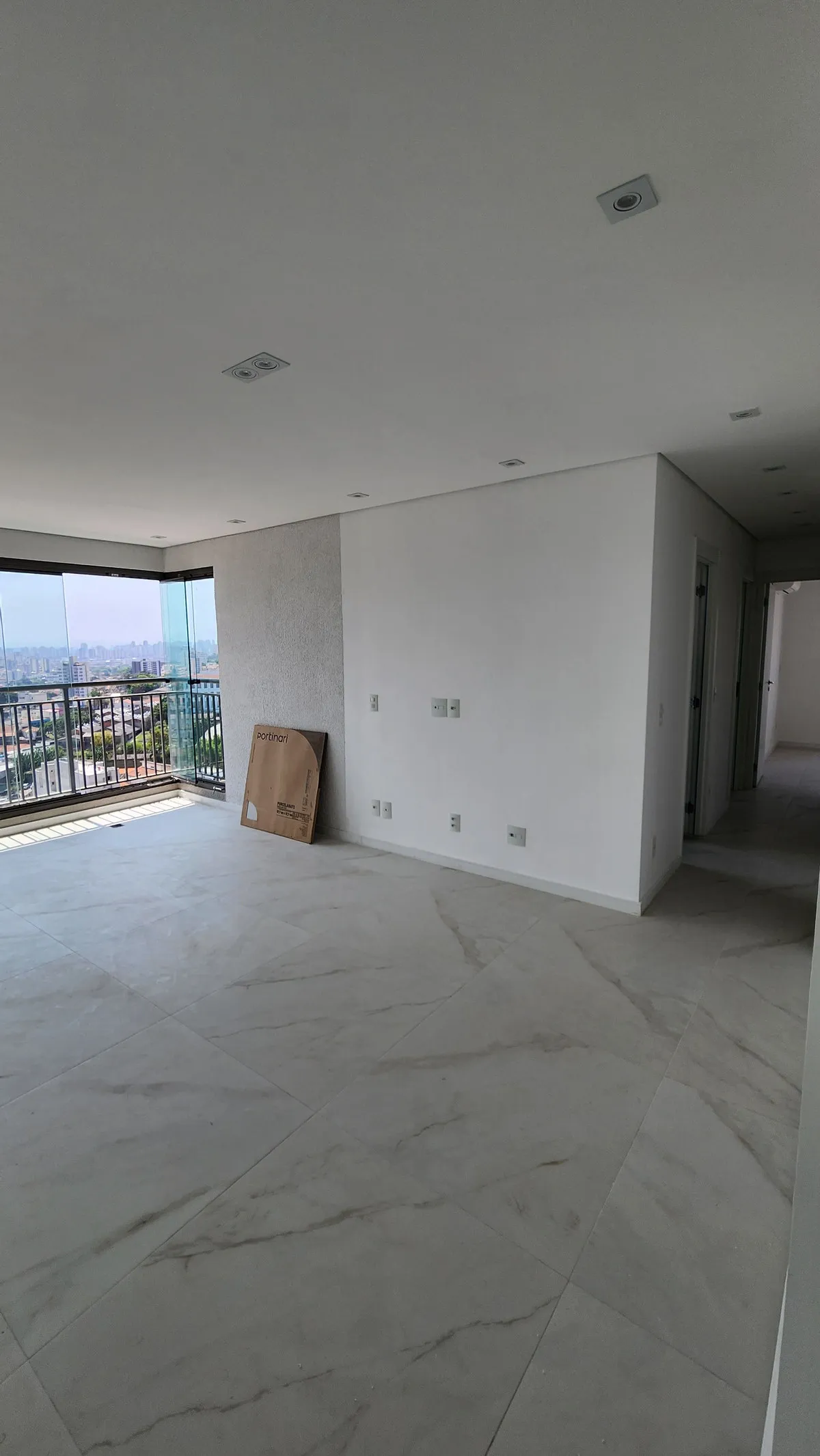 Sala vazia com piso marmorizado, parede branca e varanda envidraçada ao lado do corredor, antes da reforma