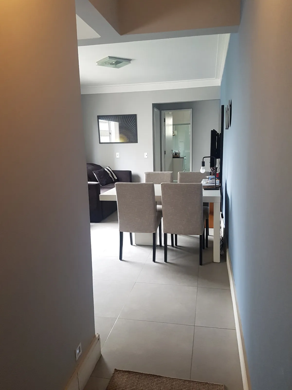 Corredor cinza abrindo para a sala de jantar com mesa branca, cadeiras estofadas cinza e sofá escuro, antes da reforma