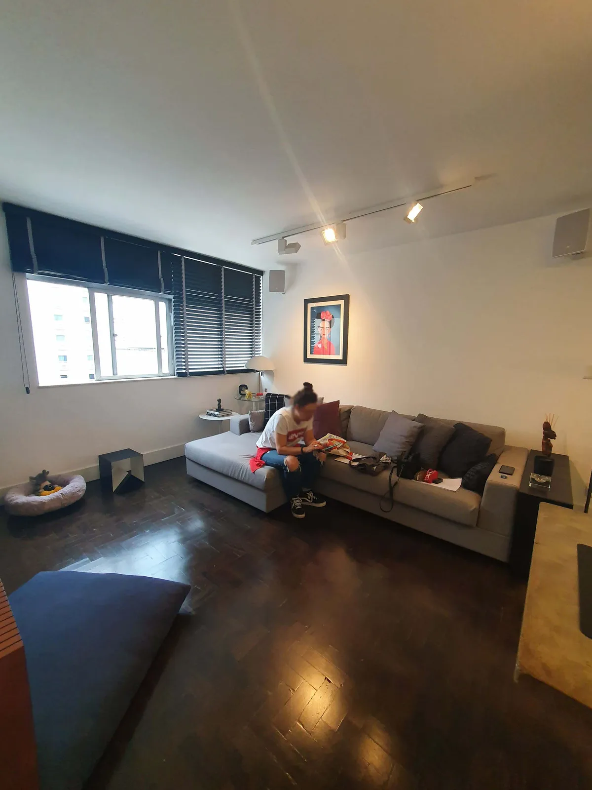 Sala de estar com sofá cinza, persiana escura na janela e piso de madeira escura, antes da reforma