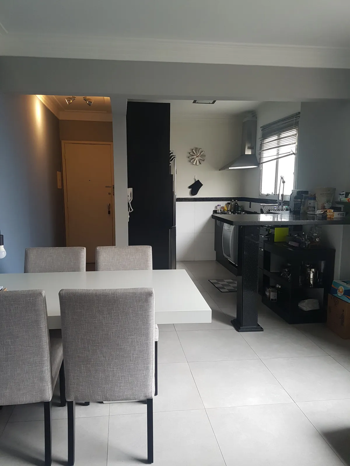 Jantar e cozinha integrados com porta de entrada bege, mesa branca, cadeiras cinza e coifa inox ao fundo, antes da reforma