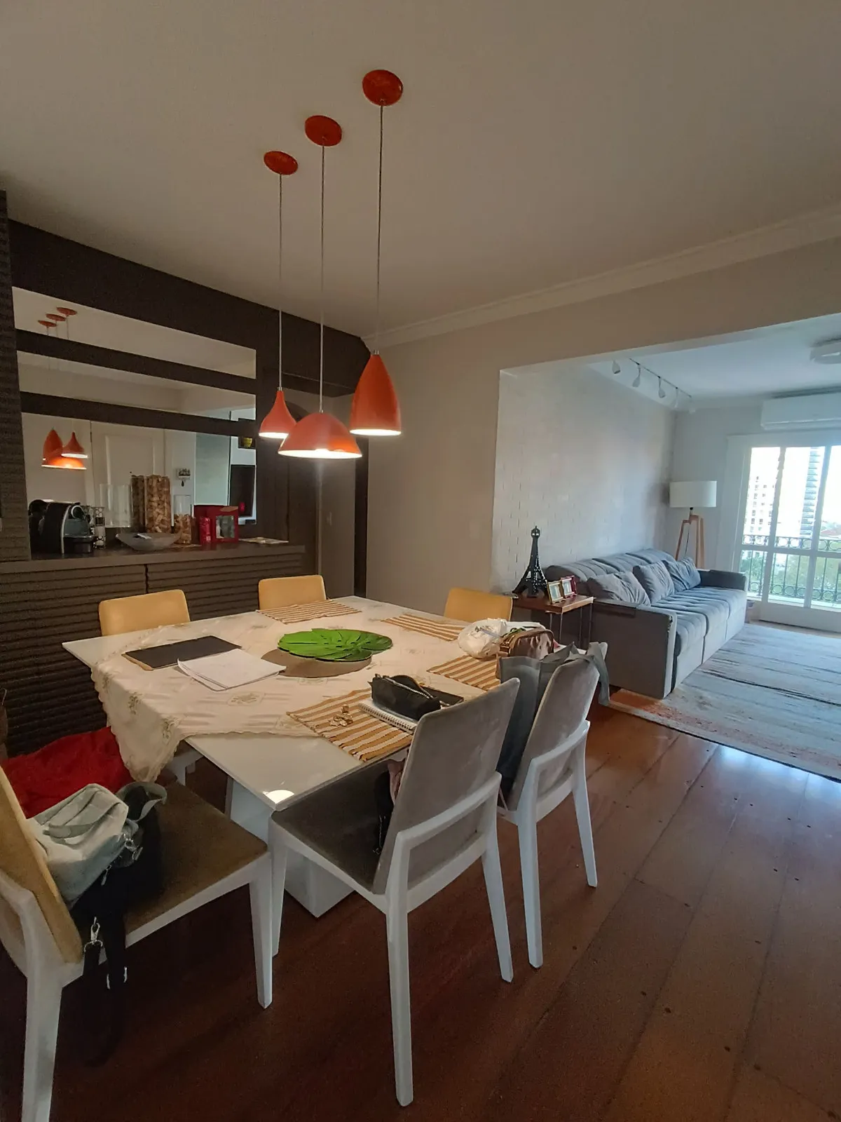 Jantar integrado à sala, com mesa branca, pendentes laranja, painel de madeira escura e sofá cinza, antes da reforma
