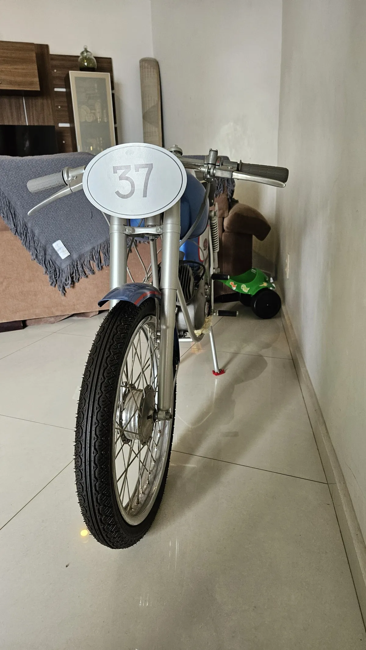 Moto antiga de corrida vista de frente, número 37, exposta na sala de piso branco, antes da reforma