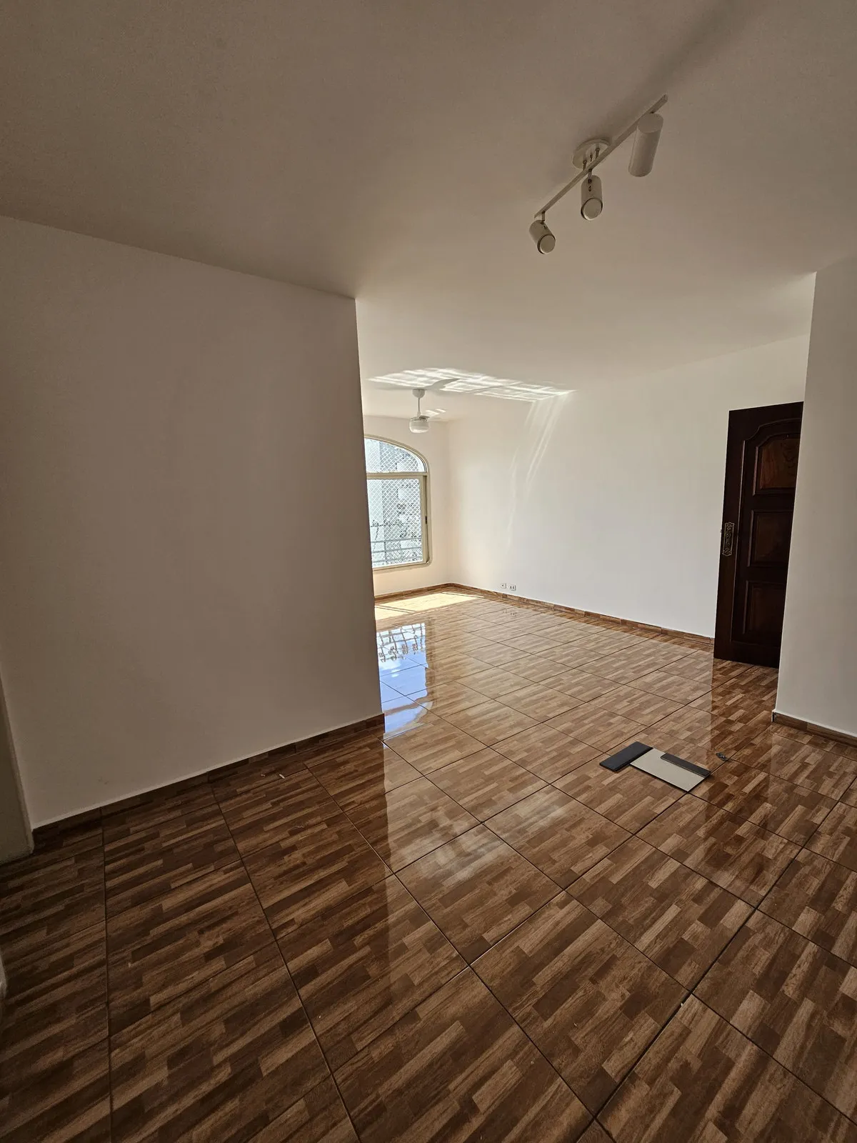 Sala vazia com piso cerâmico amadeirado, porta de madeira escura e spots de trilho no teto, antes da reforma