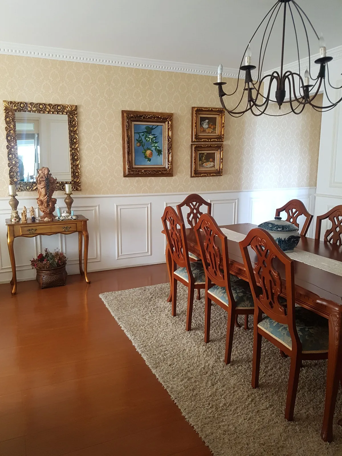 Sala de jantar com papel de parede damasco bege, aparador dourado, espelho ornamentado e mesa de madeira escura, antes da reforma