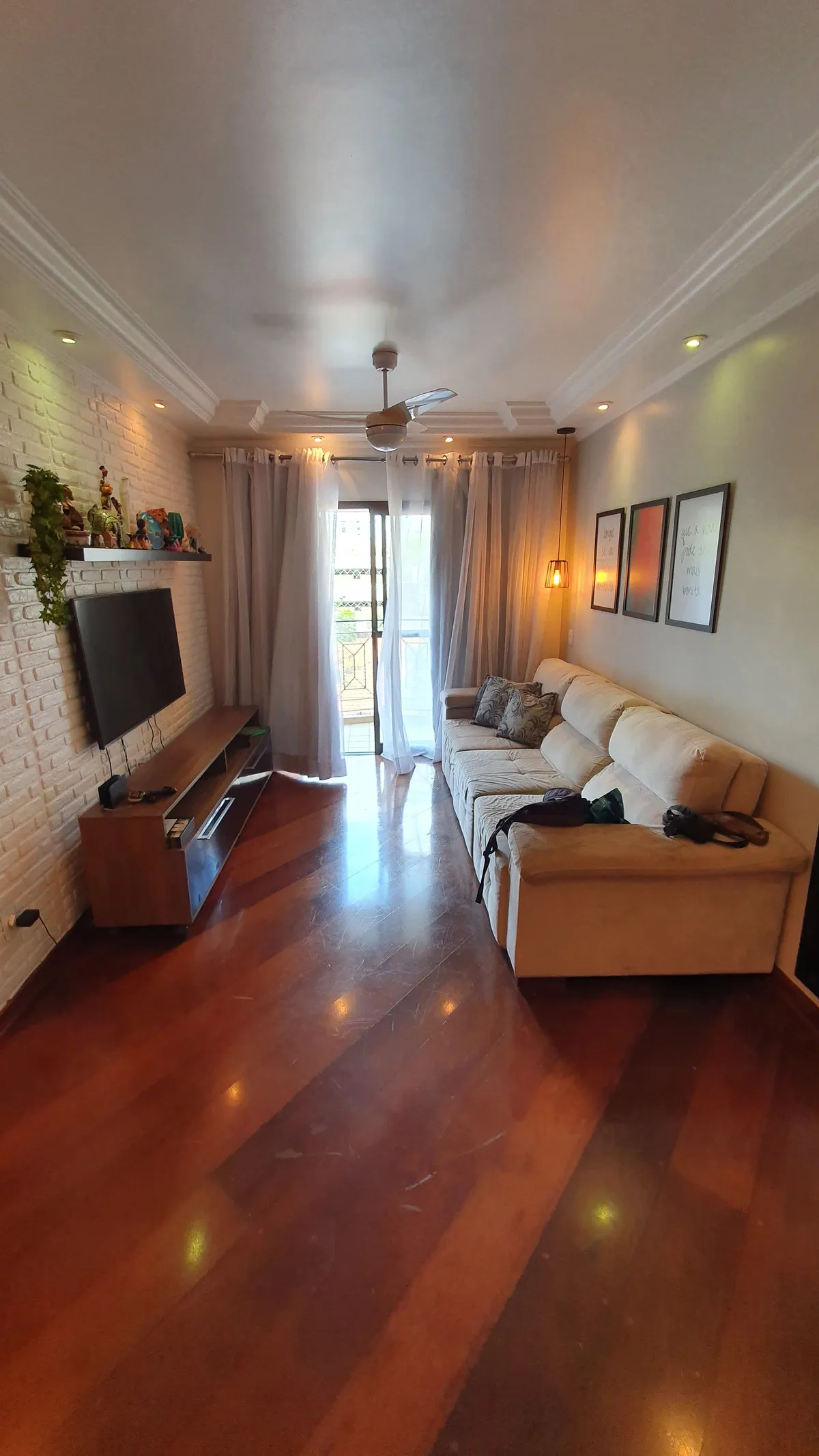 Sala de estar com TV em parede de tijolo branco, sofá bege e piso de madeira, antes da reforma