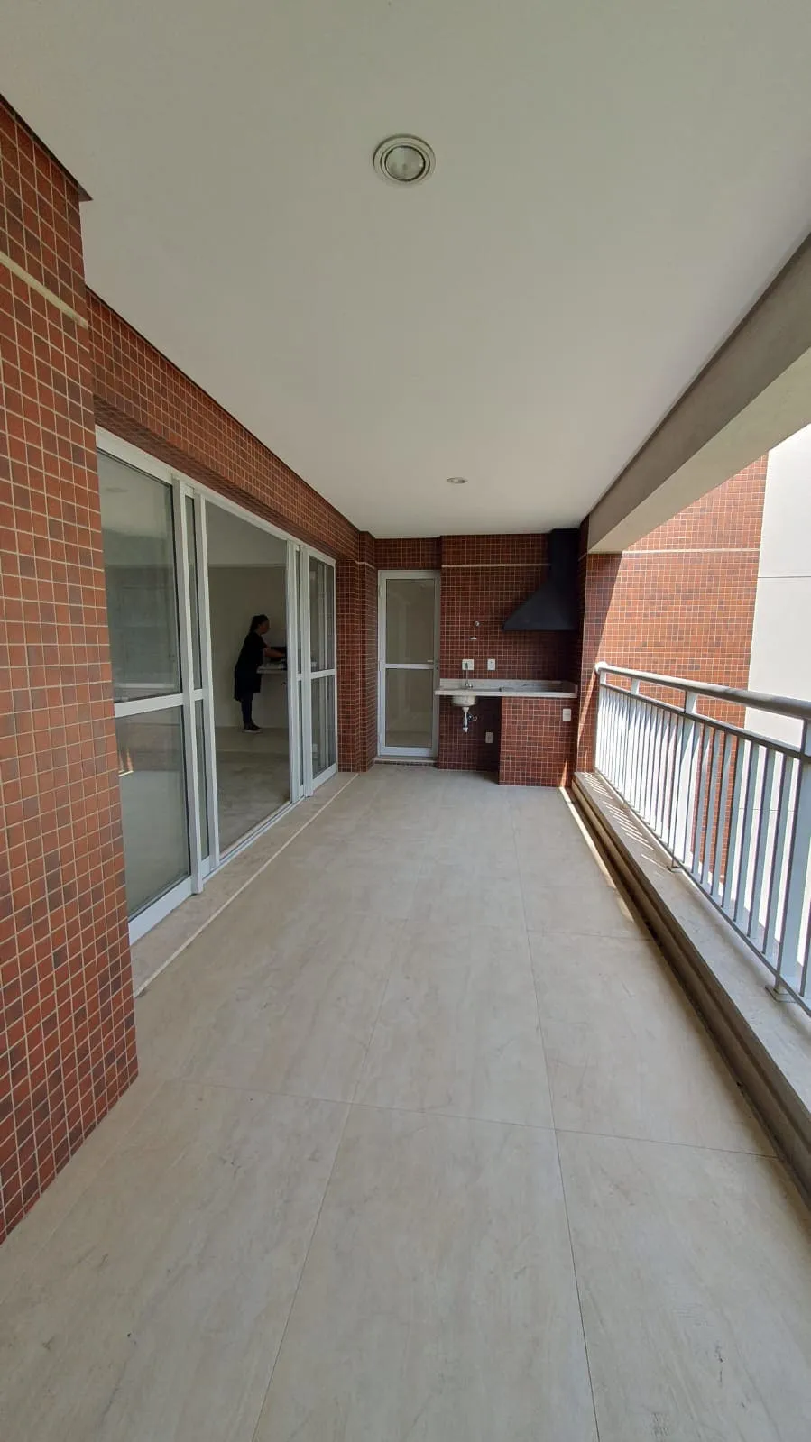Varanda com piso porcelanato claro, parede de tijolinho aparente, churrasqueira com coifa e guarda-corpo metálico, antes da reforma