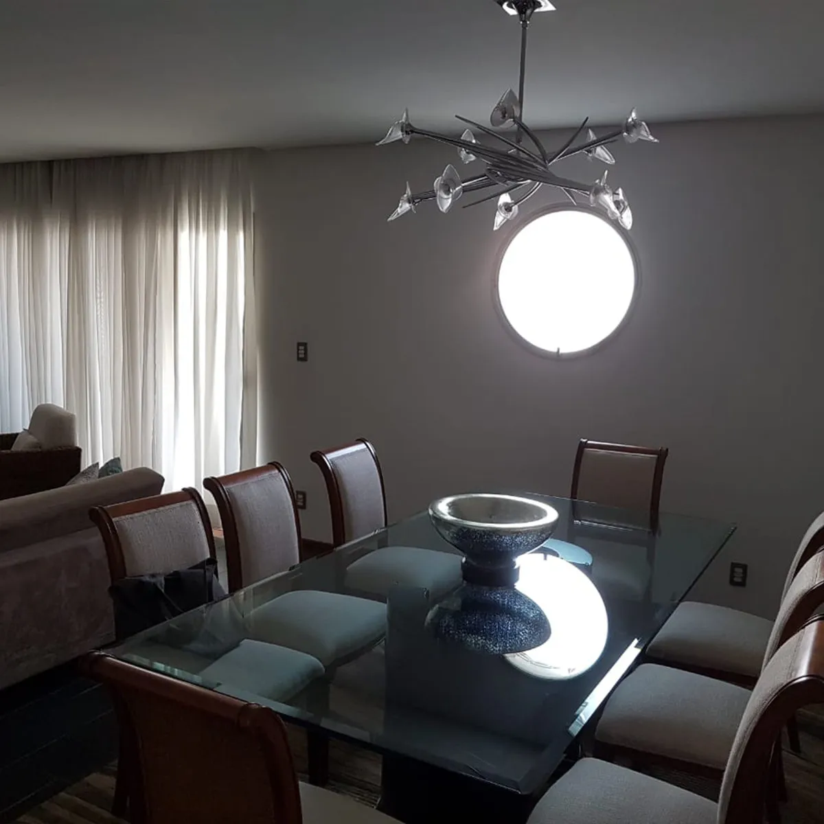 Sala de jantar escura com mesa de vidro, cadeiras de madeira, lustre e janela redonda, antes da reforma