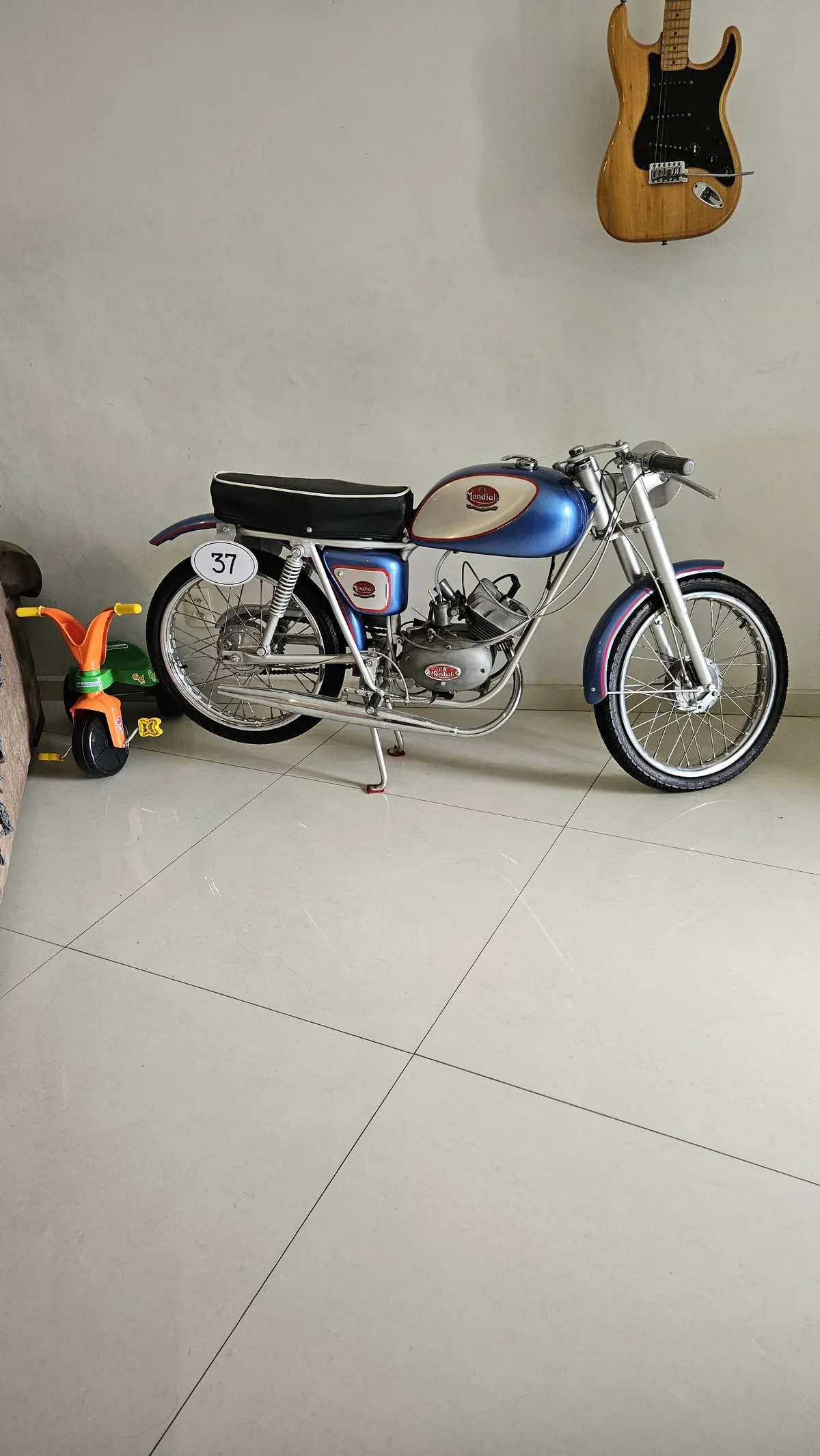 Moto Mondial azul antiga junto à parede branca, guitarra pendurada e triciclo infantil, antes da reforma