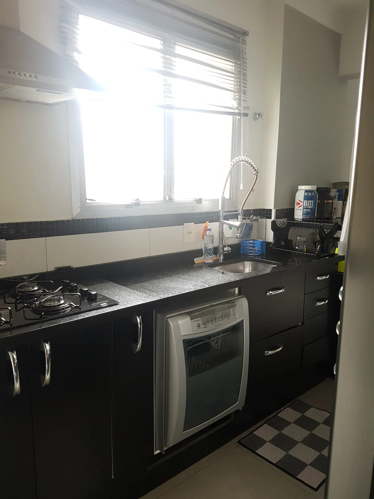 Cozinha com armários pretos, bancada de granito preto, cooktop a gás, lava-louças e janela com persiana, antes da reforma