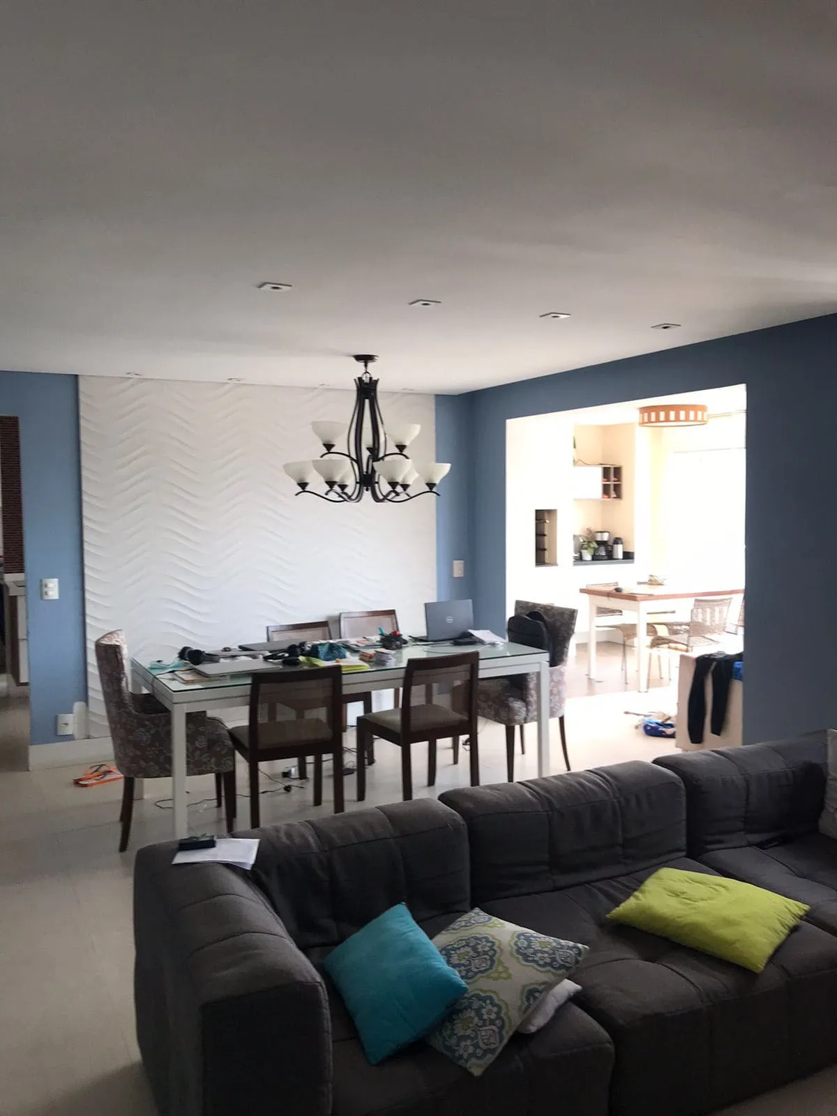 Sala de jantar com painel branco ondulado, lustre de ferro, sofá cinza e cozinha ao fundo, antes da reforma