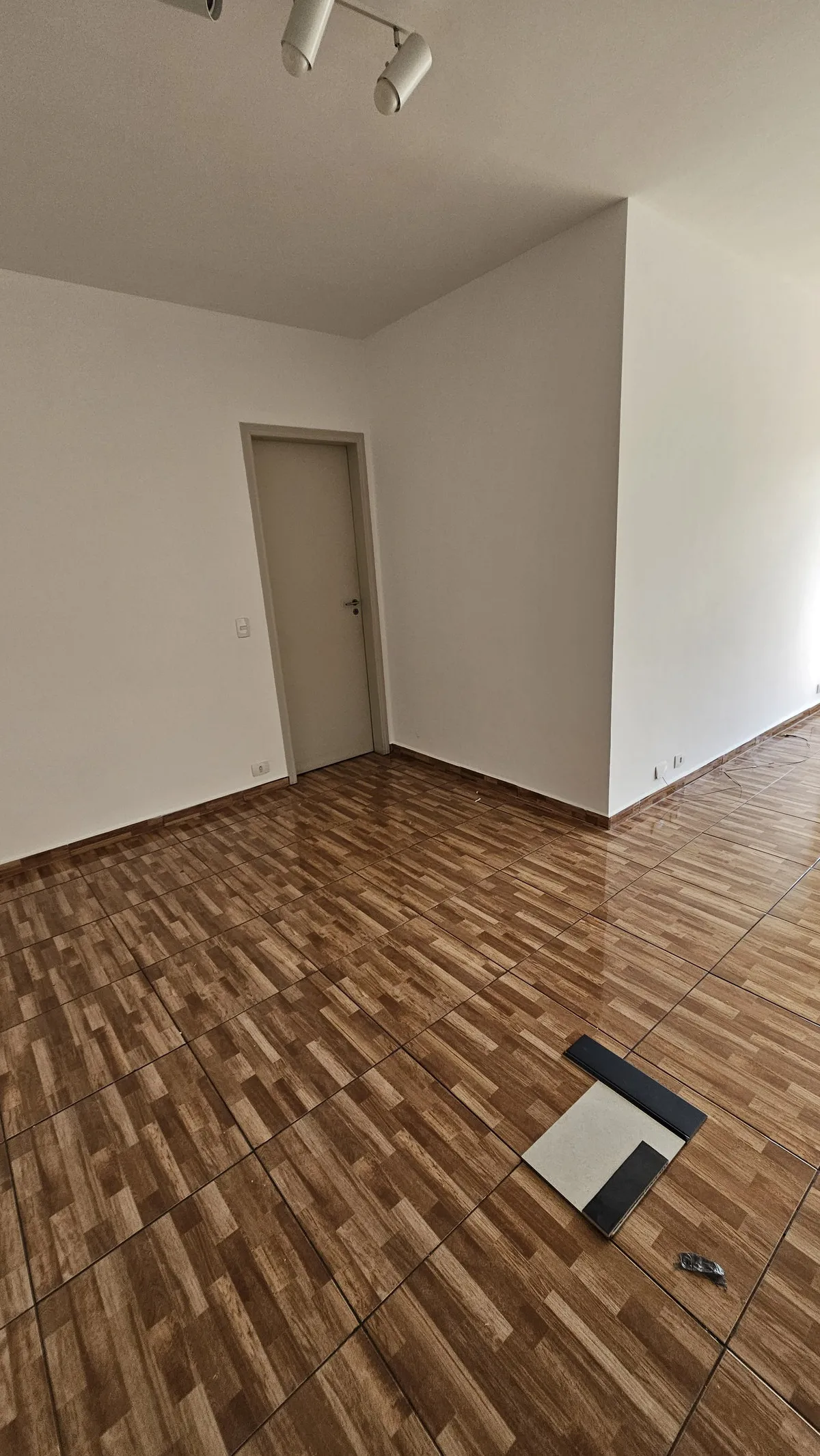 Cômodo vazio com piso cerâmico amadeirado, porta branca e trilho de spots, antes da reforma