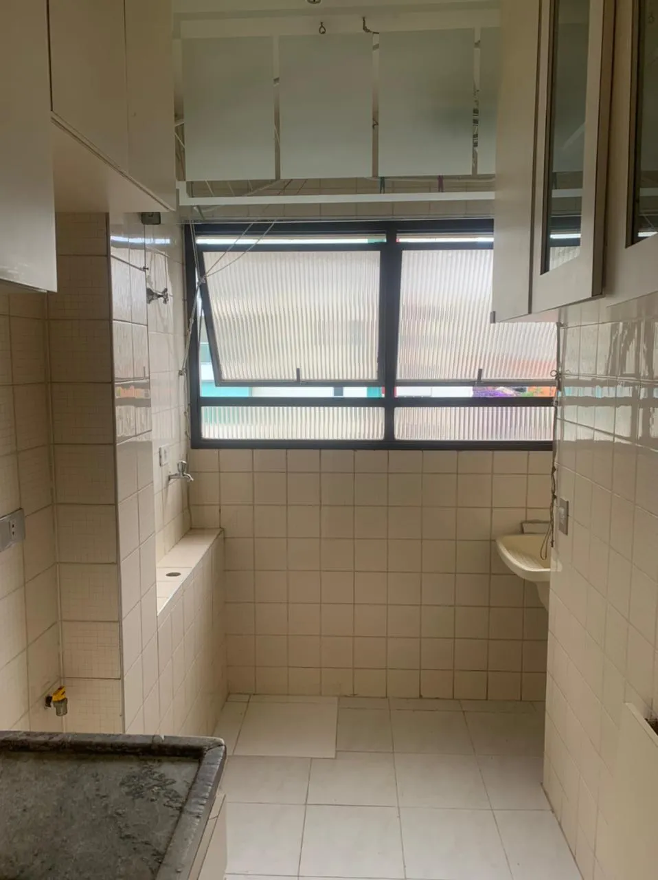 Área de serviço revestida de azulejo branco antigo, com janela basculante, tanque e armários suspensos, antes da reforma