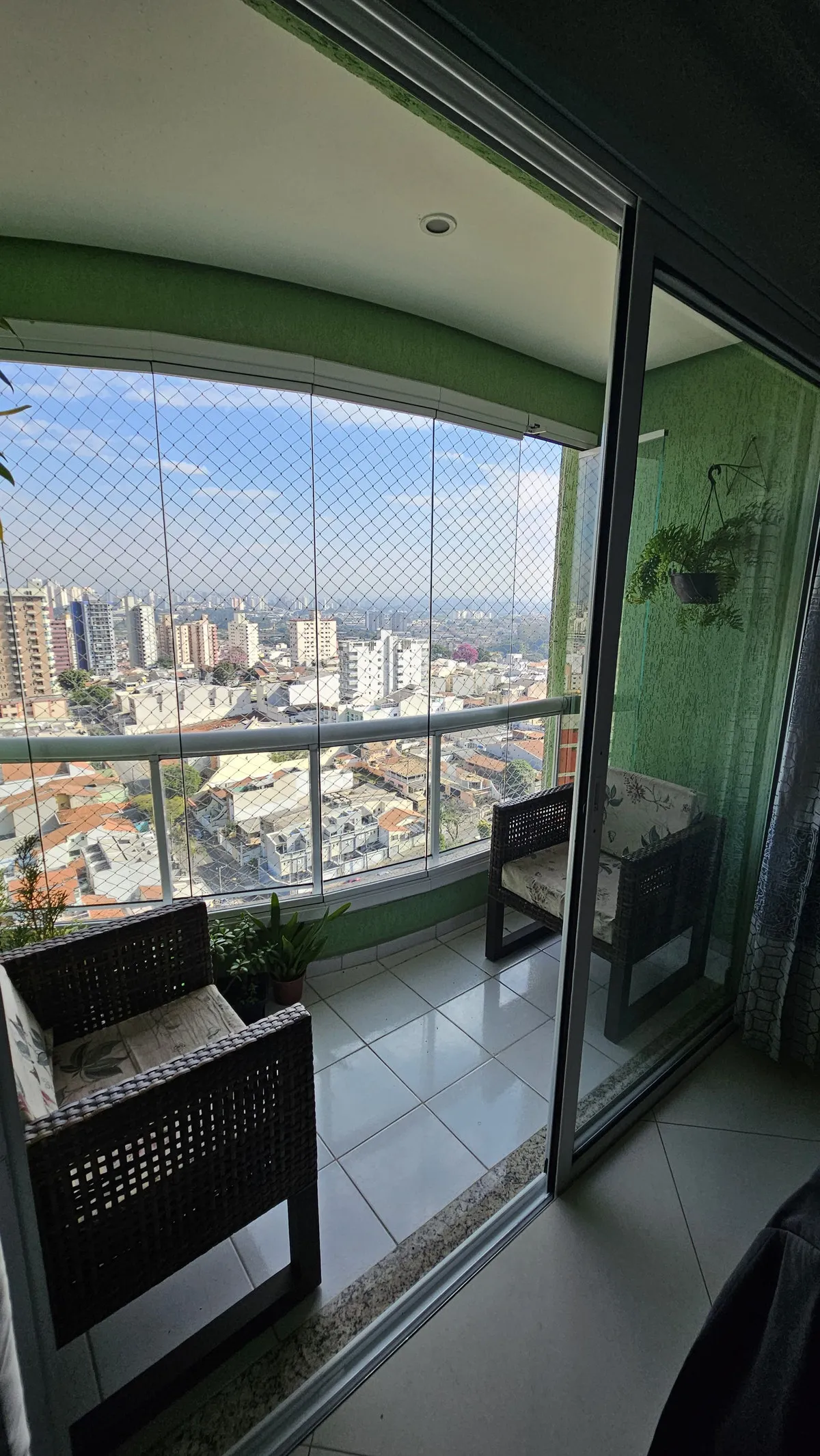 Varanda envidraçada com parede verde, cadeiras de vime e vista da cidade, antes da reforma