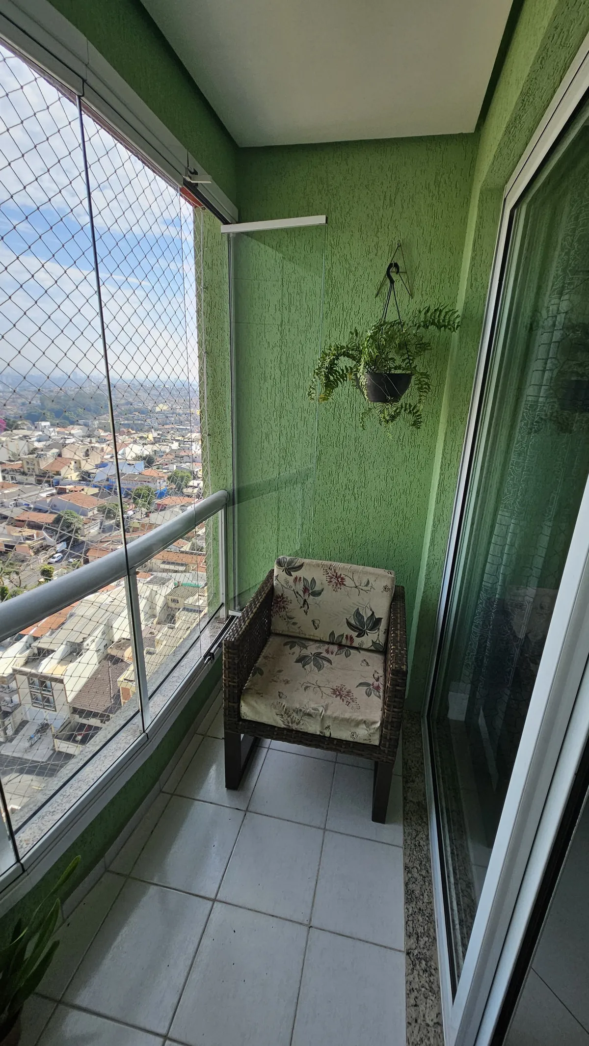 Varanda estreita com parede verde texturizada, cadeira de vime, samambaia pendurada e piso de cerâmica, antes da reforma