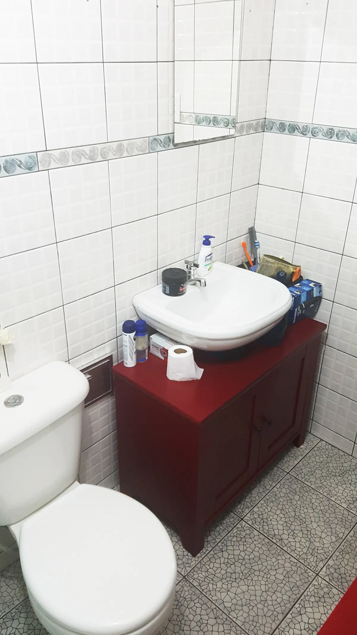 Banheiro antigo com azulejo branco de faixa decorativa, gabinete vermelho, cuba e vaso brancos, antes da reforma