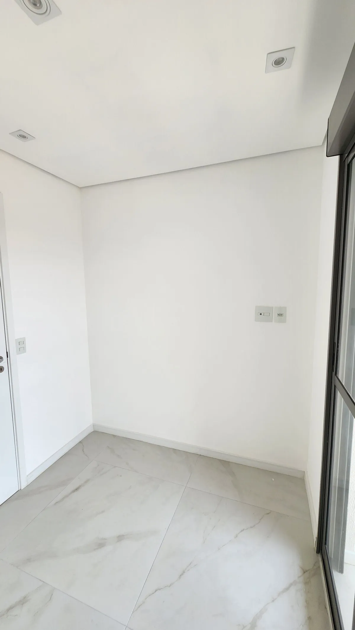Quarto pequeno e vazio com paredes brancas e porta de correr de vidro, antes da reforma