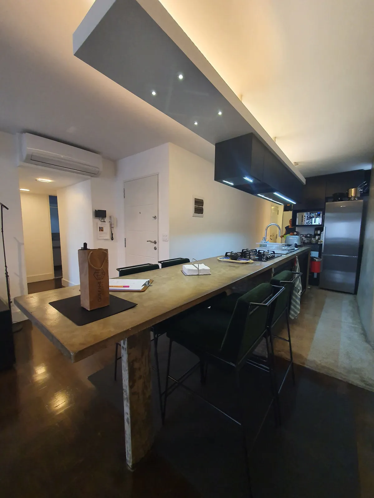 Longa bancada de concreto com banquetas pretas e geladeira inox ao fundo, antes da reforma