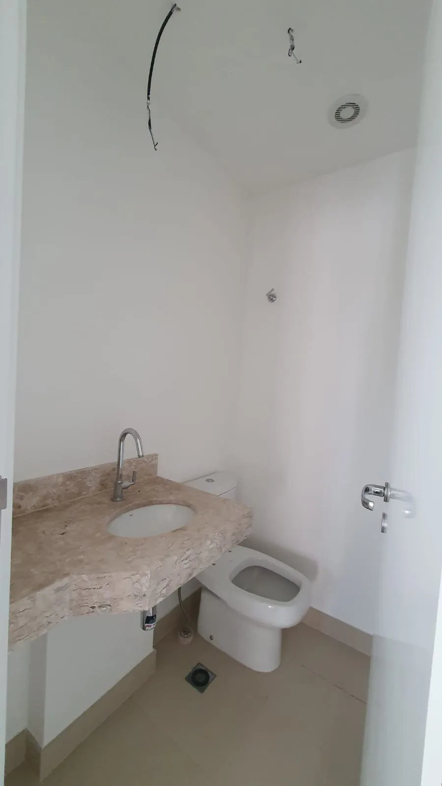 Lavabo com bancada de mármore travertino, cuba de louça, vaso sanitário e fiação aparente no teto, antes da reforma