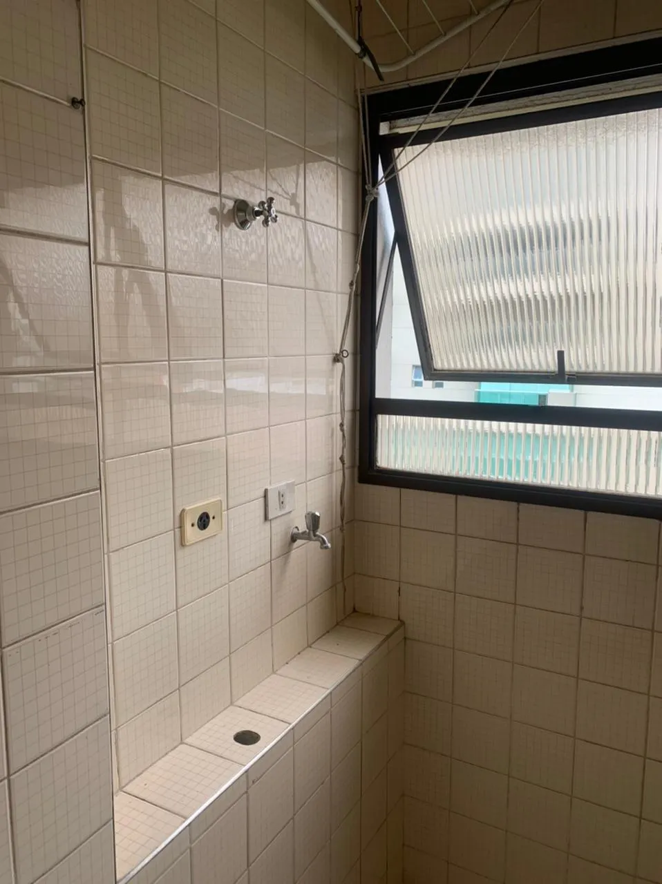 Canto de área de serviço em azulejo branco antigo, com registro, janela basculante e bancada de louça, antes da reforma