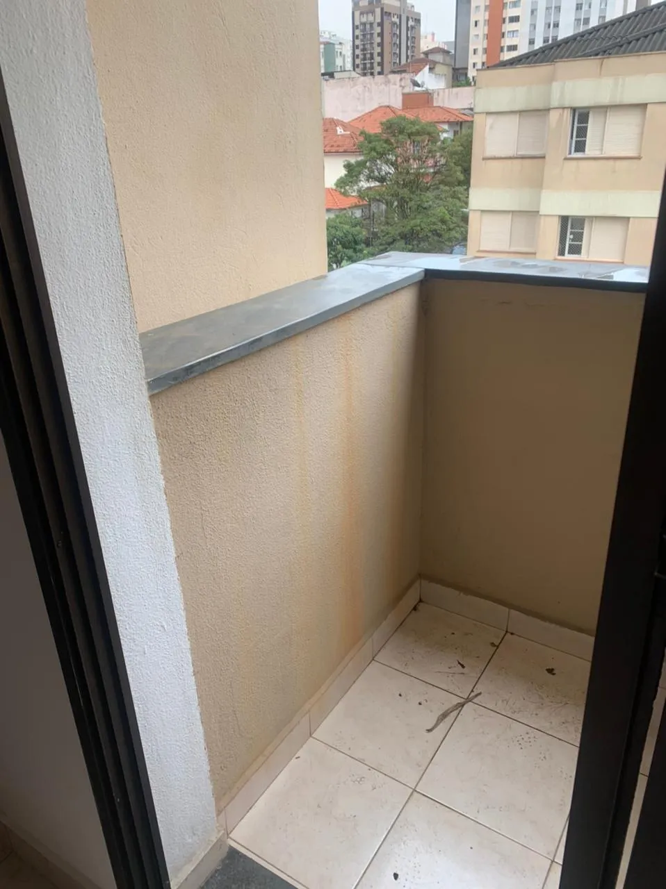 Varanda com paredes bege manchadas, piso de cerâmica e vista de prédios e casas, antes da reforma