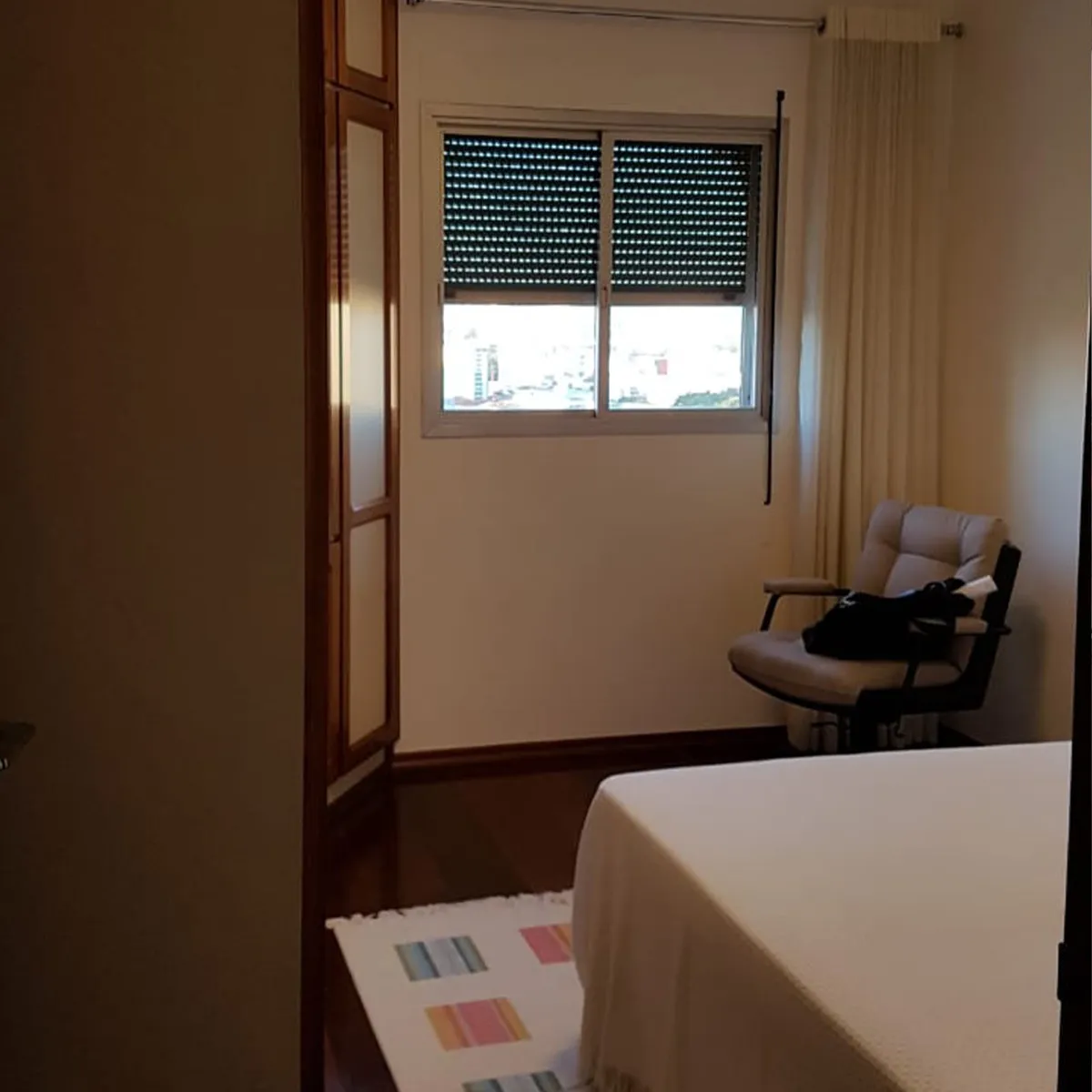 Quarto antigo com piso de taco de madeira, janela com persiana e poltrona reclinável, antes da reforma