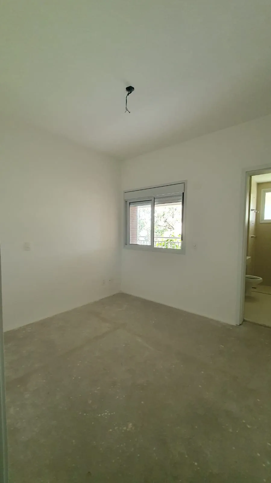 Quarto vazio com contrapiso de cimento, janela de correr e porta para banheiro à vista, antes da reforma
