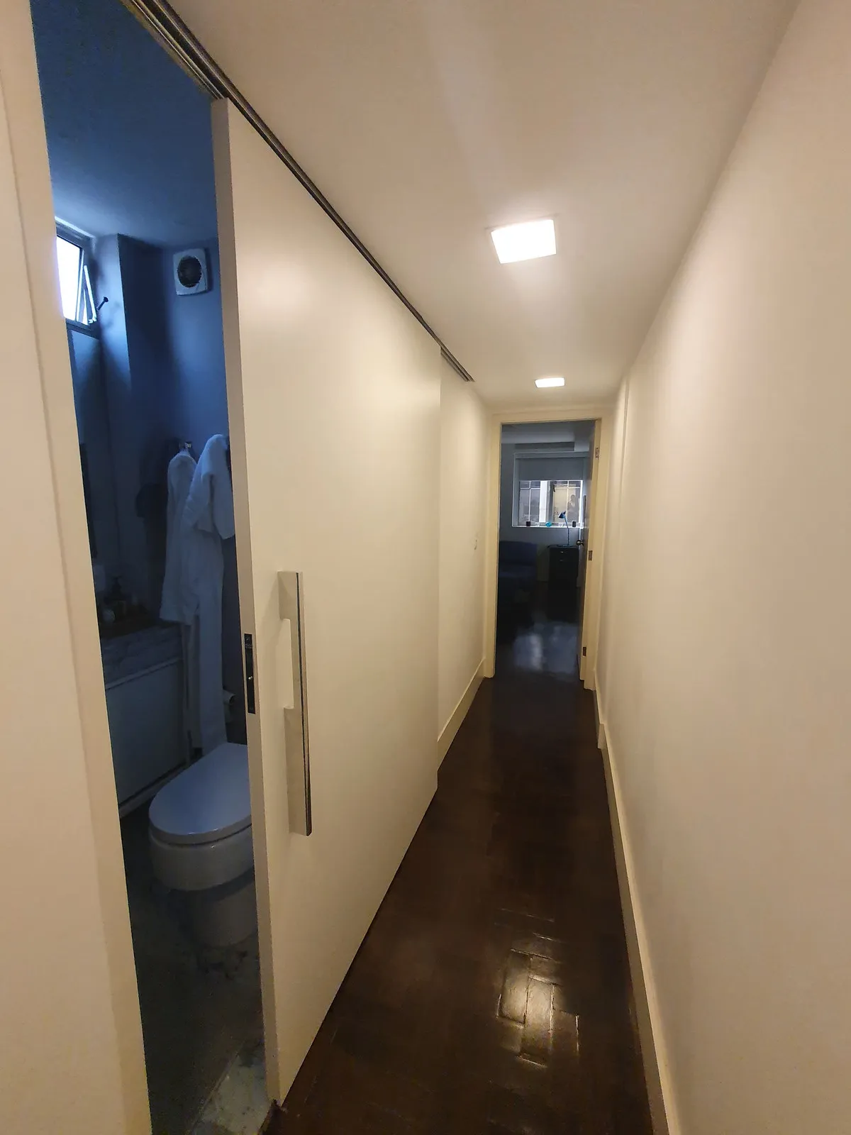Corredor estreito com porta de correr do banheiro e piso de taco escuro, antes da reforma
