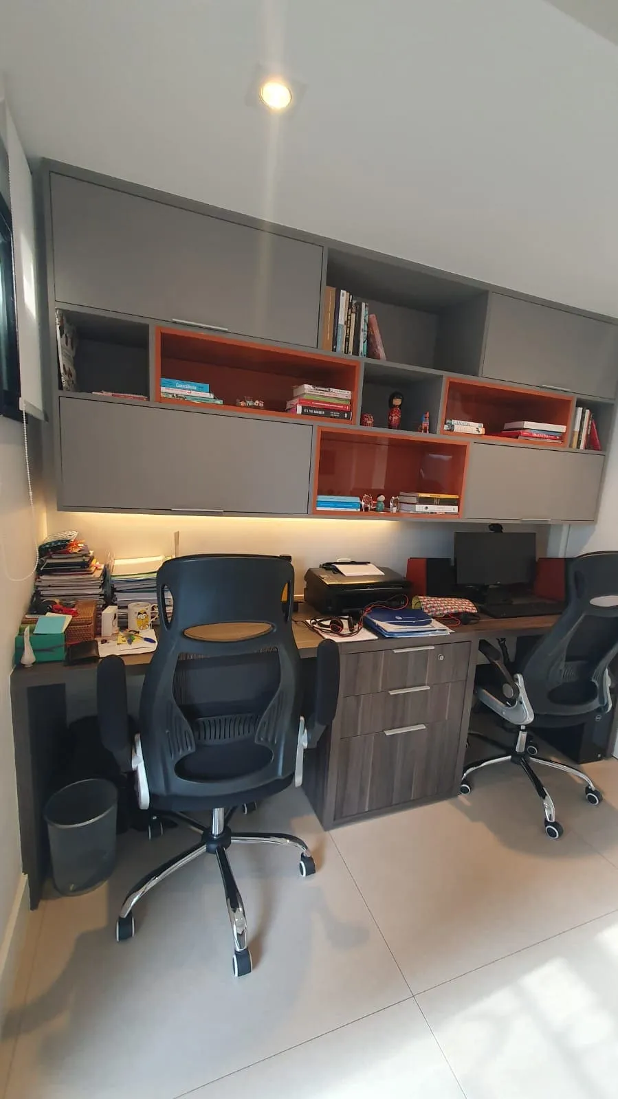 Home office antigo com armários cinza, nichos laranja cheios de livros, cadeiras pretas e bancada de madeira, antes da reforma